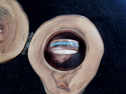 A wooden ring box with a natural wood slice lid and a circular wooden tray holding a wood ring with a turquoise inlay, set against a dark background.